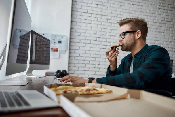 Feeling hungry. Side view of hungry male web developer in a wheelchair eating pizza and working on desktop computer while sitting at his workplace in the modern office - 写真・画像