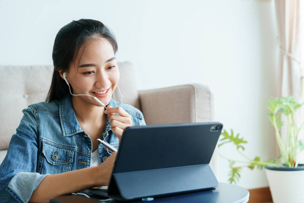 oung Asian women is conducting video conference with her co-worker using tablet computer via technology at home. beautiful woman wearing a denim shirt is talking over the headphones. work from home - 写真・画像