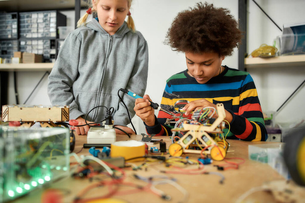Yes, we can. Boy using soldering iron to join chips and wires. His friend looking at the process. Robotics and software engineering for elementary students. Inventions and creativity for kids - Valokuva, kuva