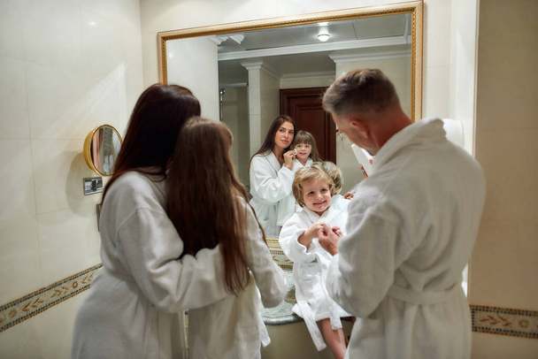 Beauty lies in a healthy mind, body, and spirit. Parents and two kids in white bathrobes having morning routine in hotel bathroom, looking in the mirror. Family, travel, resort, vacation concept - 写真・画像