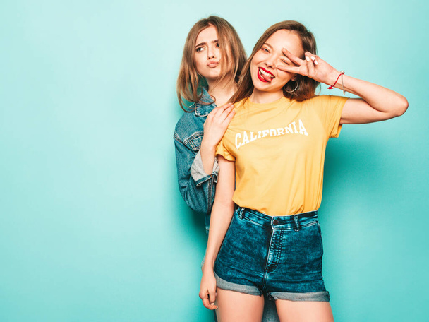 Two young beautiful smiling hipster girls in trendy summer yellow T-shirts and jeans jacket. Sexy carefree women posing near blue wall. Trendy and positive models show peace sign - Fotoğraf, Görsel