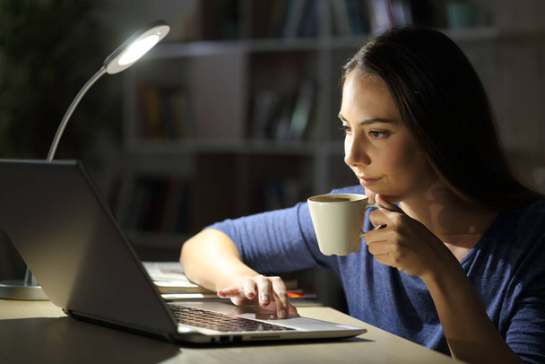 Serious woman using laptop holding coffee cup sitting alone with a desk lamp on the table at night at home - Фото, изображение