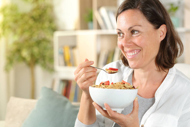 Счастливая взрослая женщина, держа в руках шейную миску, смотрит в сторону, завтракая сидя на диване в гостиной дома
 - Фото, изображение