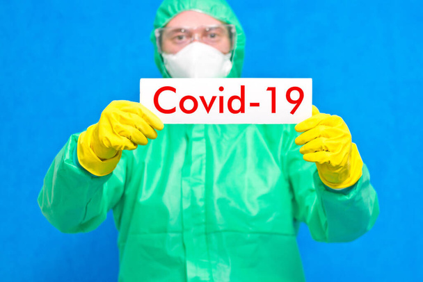 Ιατρός που κρατά λευκή αφίσα με την επιγραφή COVID-19 σε μπλε φόντο - Φωτογραφία, εικόνα