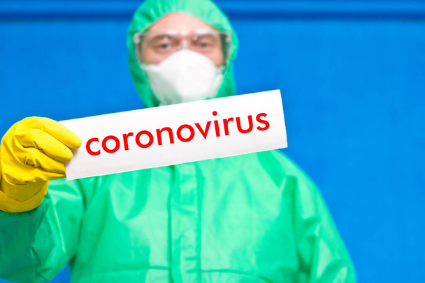 Travailleur médical tenant une affiche blanche avec l'inscription coronavirus, debout sur un fond bleu
. - Photo, image
