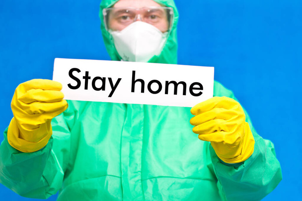 Medische medewerker met een witte poster met de inscriptie thuis blijven, staan op een blauwe achtergrond - Foto, afbeelding