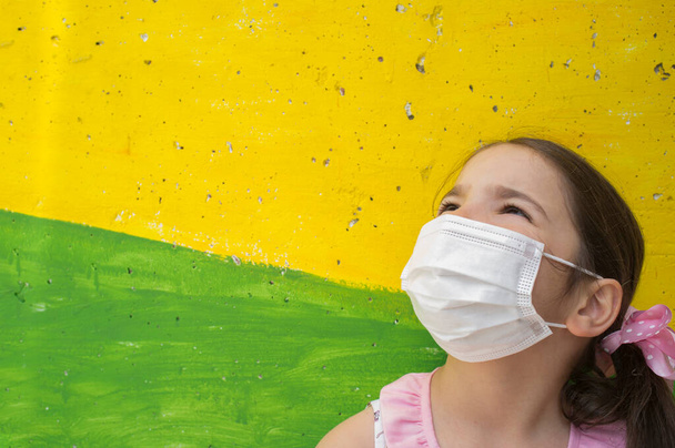 Hoffnungsvolles kleines Mädchen trägt Gesichtsmaske während der Covid-19-Pandemie. Lebendig gefärbter Betonblock als Hintergrund - Foto, Bild