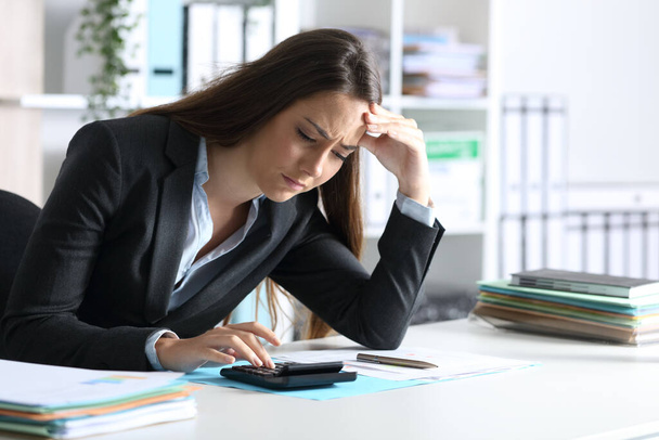 Занепокоєна жінка-бухгалтер, яка розраховує на калькулятор, сидячи на столі в офісі
 - Фото, зображення