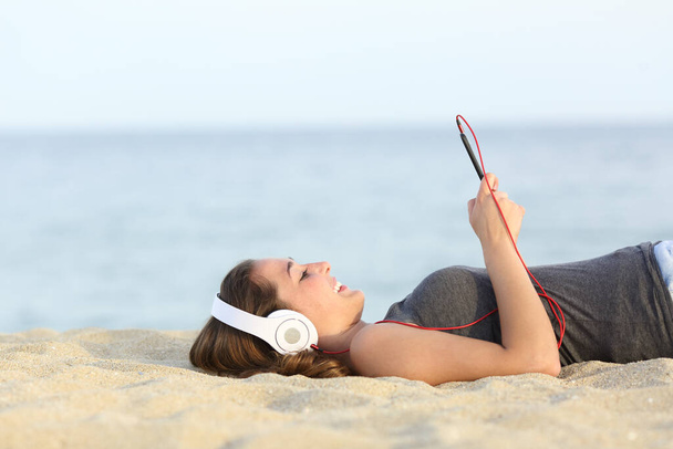 Side view of a happy girl listens music on smart phone with headphones lying on the sand on a beach - Fotó, kép