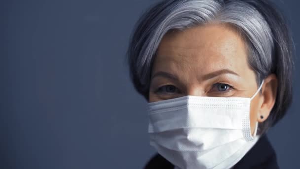 White-haired business woman in a protective mask looks at the camera standing on a dark background. Business concept. Close up. Prores 422 - Materiał filmowy, wideo