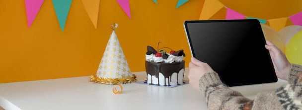 Обрізаний знімок жінки, що тримає цифровий планшет, сидячи за столом з тортом і партійним капелюхом
   - Фото, зображення
