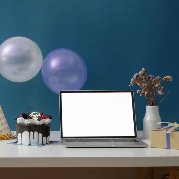 Online verjaardagsfeest concept met blanco scherm laptop, taart, feestmuts en decoraties op witte tafel met blauwe wand - Foto, afbeelding