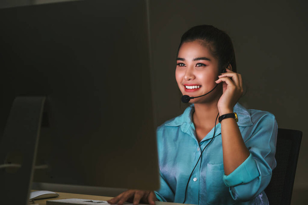 Servicio de atención al cliente femenino asiático sonriendo y trabajando duro hasta tarde en el turno de noche en la oficina, departamento de centro de llamadas, trabajador y horas extras, trabajo en equipo con colegas para el concepto de éxito
 - Foto, Imagen