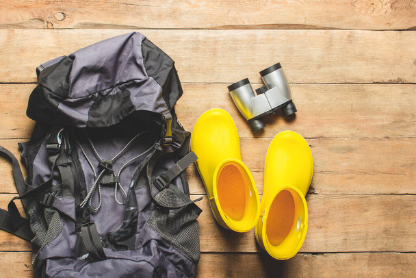 Yellow rain boots, backpack, binoculars, camping equipment on a wooden background. The concept of hiking, tourism, camp, mountains, forest. Banner. Flat lay, top view. - Photo, Image