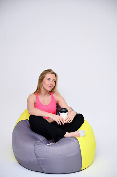 Portrait of a pretty fitness blonde girl in a sports uniform on a white background sits in a soft chair with a cup of coffee in her hands. A slender Caucasian model poses with a smile. - Foto, immagini