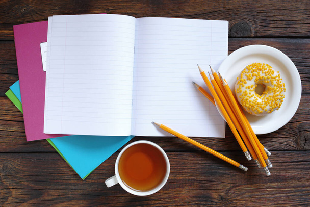 Iskolai jegyzetfüzet ceruzával, csésze tea és fánk fából készült asztalon, felülnézet - Fotó, kép