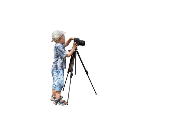 Ένα μικρό αγόρι τραβάει φωτογραφίες με μια κάμερα σε ένα τρίποδο σε λευκό απομονωμένο φόντο - Φωτογραφία, εικόνα