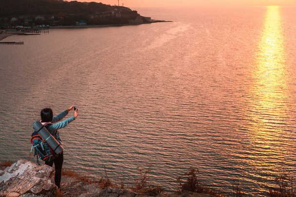 Une femme se tient au bord d'une falaise et prend des photos du coucher de soleil sur son smartphone. Au fond, la mer au coucher du soleil. Espace de copie. Concept de randonnée et d'activités de plein air
. - Photo, image