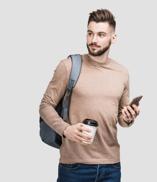 Молодий красивий чоловік з рюкзаком тримає смартфон і каву ізольовано на сірому фоні. Серйозний студентський або бізнесмен студійний портрет. Веселі чоловіки вирушають у подорож
. - Фото, зображення