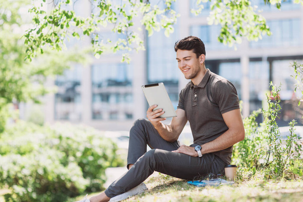 Smiling man student using digital tablet in a city park. Young handsome men having coffee break. Modern lifestyle, connection, business, freelance work, education concepts - Foto, afbeelding