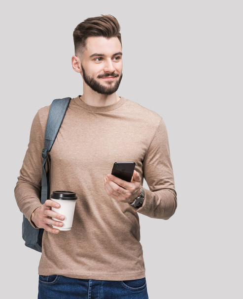 Young handsome man with backpack holding smart phone and coffee isolated on gray background. Serious student or businessman studio portrait. Cheerful men going on a travel. - Фото, зображення