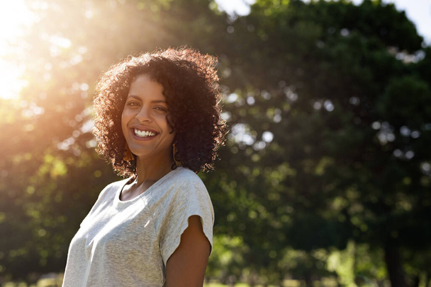 Portrait of a young woman with curly hair smiling while standing outside in a park on a sunny summer afternoon - Foto, Imagen