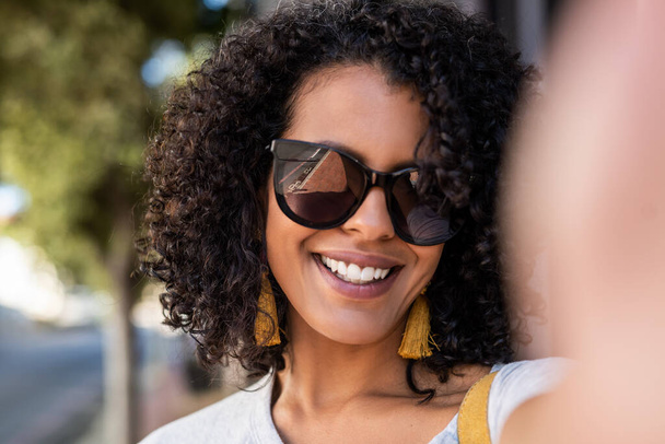Smiling young woman with curly hair and wearing sunglasses taking a selfie while standing on a sidewalk in the city - Fotografie, Obrázek