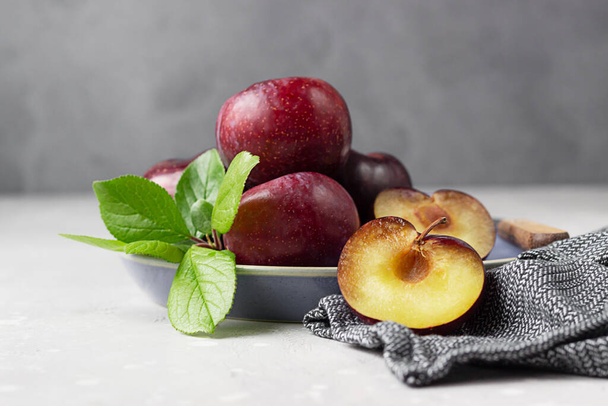 Whole purple plums and slices with leaves and knife on plate, light grey concrete background. - Fotoğraf, Görsel