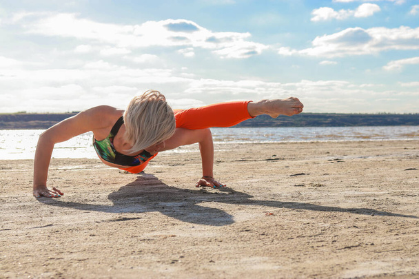 Junge blonde Frau in roten Leggings, die im Sommer Morgengymnastik an der Küste macht. Fitness Yoga Stretching Training draußen. Gesundes Leben. Aktive Lebensweise - Foto, Bild