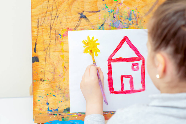 Рука ребенка рисует желтое солнце и красный дом с акварелью на листе бумаги дома. Девочка рисует в школе
. - Фото, изображение