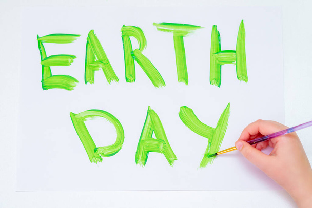Draufsicht der Hand eines Kindes, das Wörter Earth Day mit Pinsel von grünem Aquarell auf weißem Papier schreibt. Konzept des Tages der Erde. - Foto, Bild