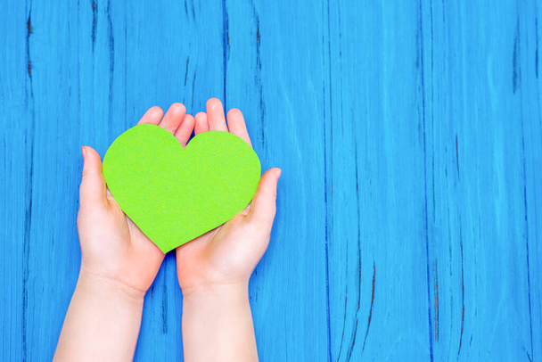 Top view of hands of child holding green heart on wooden blue background. Copy space. Concept of environmental protection. - 写真・画像