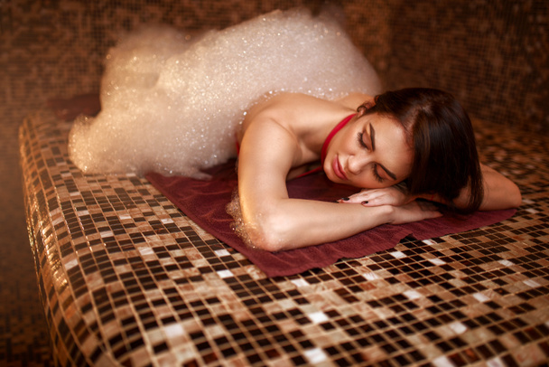 Frau in Schaumstoff auf heißem Stein liegend, türkisches Bad, Hamam, Sauna, Draufsicht. Haut- und Körperpflege - Foto, Bild