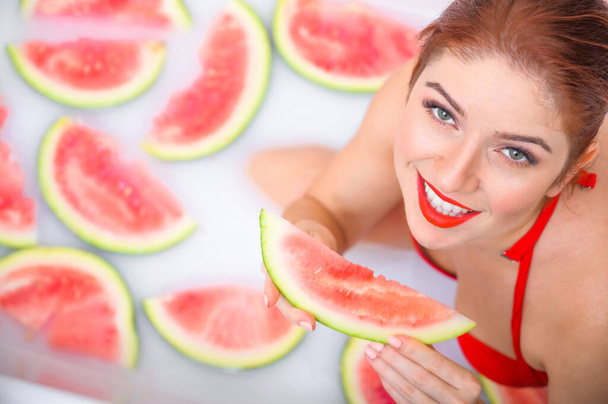 Portrait of a woman in a red swimsuit eating a watermelon and smirking. Redhead girl with red lipstick takes a bath with milk and fruit to rejuvenate her skin. Body care. - Fotografie, Obrázek
