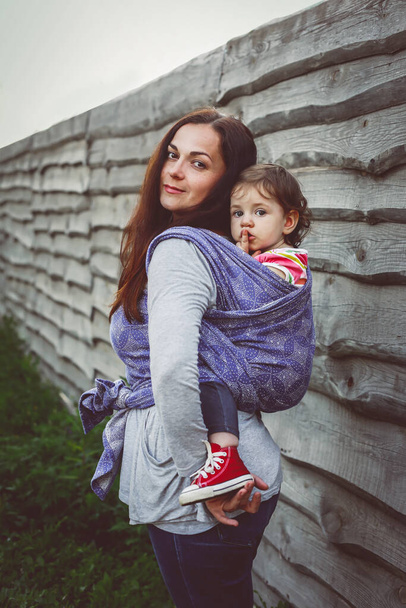 Mère tenir bébé fille dos dans le porte-bandoulière à fond de clôture en bois rural, mignon enfant femelle et maman moderne active
 - Photo, image