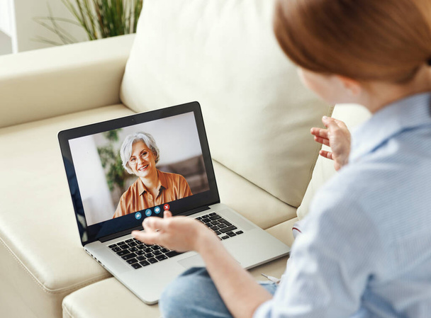 Young woman   enjoying video conversation via laptop with eldery mother while sitting on sofa  - Fotó, kép