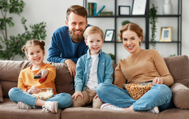 Gelukkig jonge ouders met schattige kinderen zitten op de bank in de woonkamer thuis en kijken grappige film terwijl het eten van popcorn en genieten van weekend samen - Foto, afbeelding