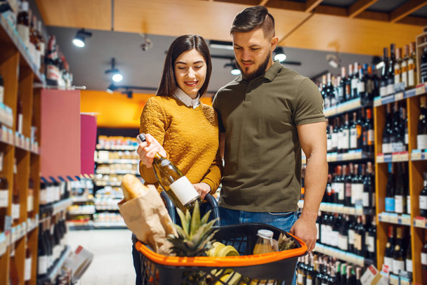 Atractiva pareja que elige el vino en el supermercado de comestibles. Hombre y mujer con carro comprando bebidas en el mercado, clientes comprando alimentos y bebidas
 - Foto, imagen