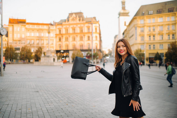 A young beautiful stylish girl in a black summer dress and jacket and with a purse walks through the old city.Girl-model in the Czech Republic. BRNO. - 写真・画像