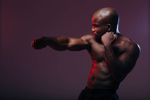 Um pugilista de pele escura está a treinar com um tronco nu. O atleta com músculos em relevo é retroiluminado com luz vermelha
. - Foto, Imagem