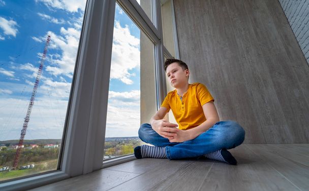 Boy sitting at floor at balcony. Kid looking at nature outside window. isolation at home while quarantine. Sad boy dreaming. - Foto, Imagen