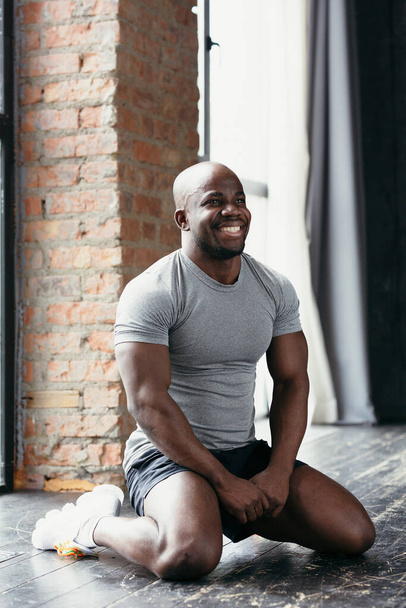 Portrait of a black pilates trainer in a gray t-shirt sitting on his knees on the floor in the gym and laughing - Foto, Imagem