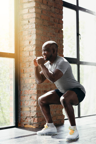 Profile view Athletic African American in a tight T-shirt doing squats while holding his arms near his chest in the gym - Фото, изображение