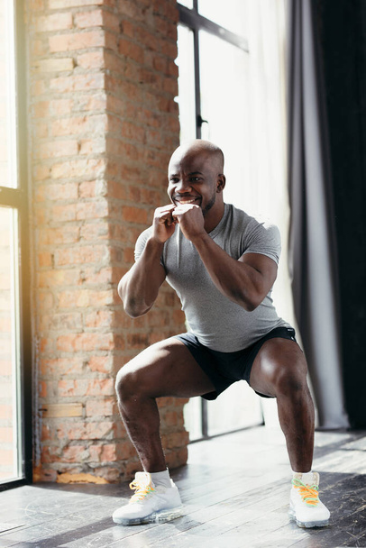 Athletic African American in a tight-fitting T-shirt does squats while holding his hands near his chest in the gym - Φωτογραφία, εικόνα