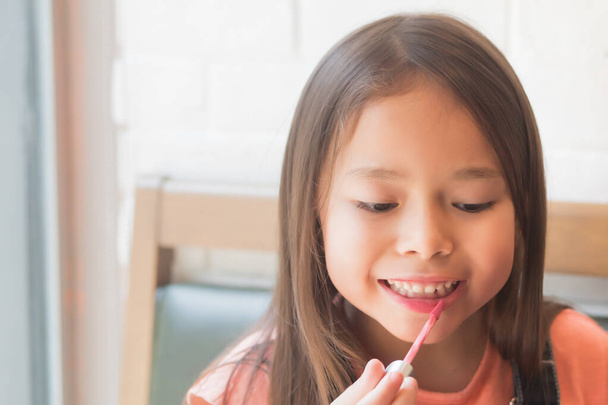 cute little girl playing and applying makeup lipstick to her lips; concept of girl grooming, face care, beauty face, kid playing with cosmetics and lipstick; asian caucasian kid girl model - 写真・画像