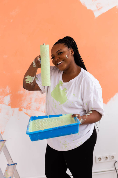 Νεαρή όμορφη Αφροαμερικανή ζωγραφίζει τοίχο στο νέο της διαμέρισμα. Σχέδιο ανακαίνισης και ανακαίνισης. - Φωτογραφία, εικόνα