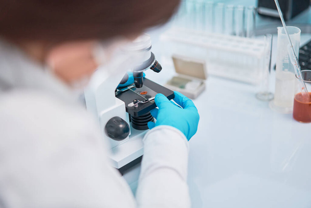 Κοντινό πλάνο της γυναίκας βοηθός εργαστηρίου σε μπλε λαστιχένια γάντια κοιτάζοντας μέσα από μικροσκόπιο - Φωτογραφία, εικόνα