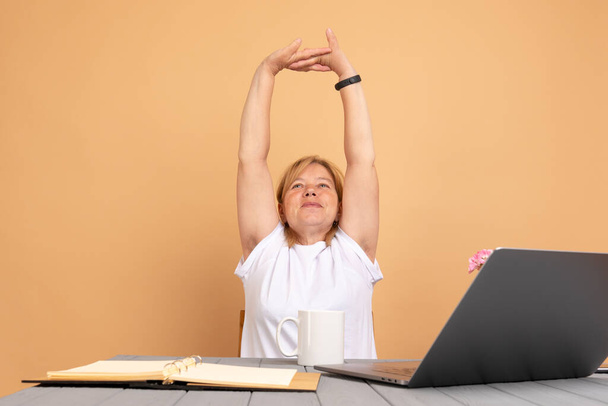 Ανώτερη γυναίκα κάθεται στο τραπέζι με φορητό υπολογιστή, αναπαύεται με υψωμένα χέρια, δείχνοντας τριχωτές αξύριστες γυναικείες μασχάλες. Θετική τάση σώματος - Φωτογραφία, εικόνα