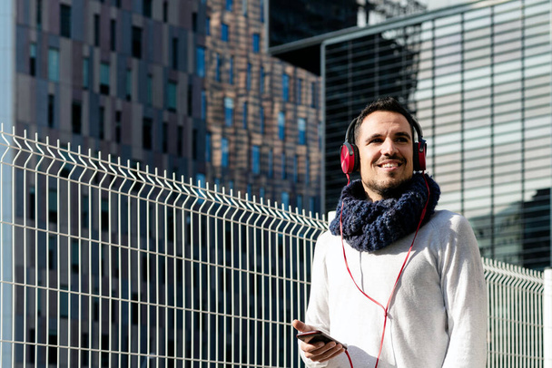 Young bearded man with headphones and holding smartphone while walking against skyscrapers in sunny day - Foto, Imagen