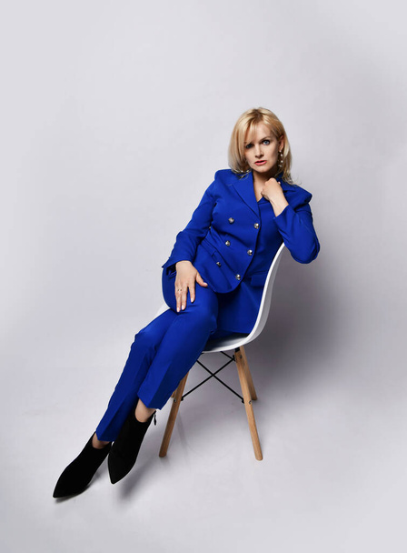 Powerful blonde woman in stylish deep blue official costume and shoes is sitting on a chair looking at us - Foto, Imagem